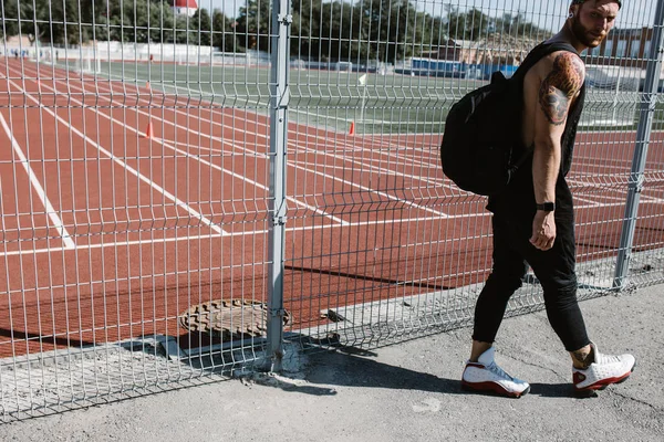 Homme athlétique en bandeau vêtu de vêtements de sport noirs avec sac à dos sur les épaules marche le long de la clôture de l'aire de jeux par une journée ensoleillée — Photo