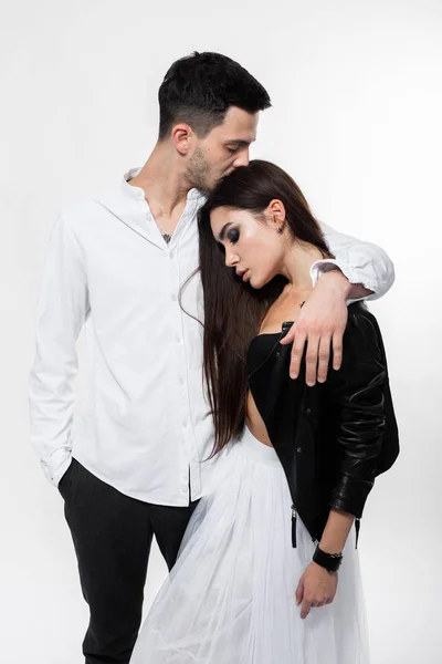 Menina morena bonita em uma saia de tule branco em um sutiã preto e jaqueta de couro preto com cara de calças e camiseta branca estão abraçando no fundo branco no estúdio — Fotografia de Stock