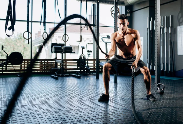 Homem de cabelos escuros atlético com um tronco nu vestido com shorts pretos fazendo exercícios esportivos com cordas esportivas no ginásio — Fotografia de Stock