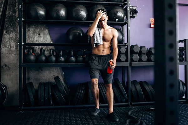 Homem atlético com um tronco nu e t-shirt em seu ombro vestido com os shorts pretos fica perto do equipamento esportivo no ginásio — Fotografia de Stock