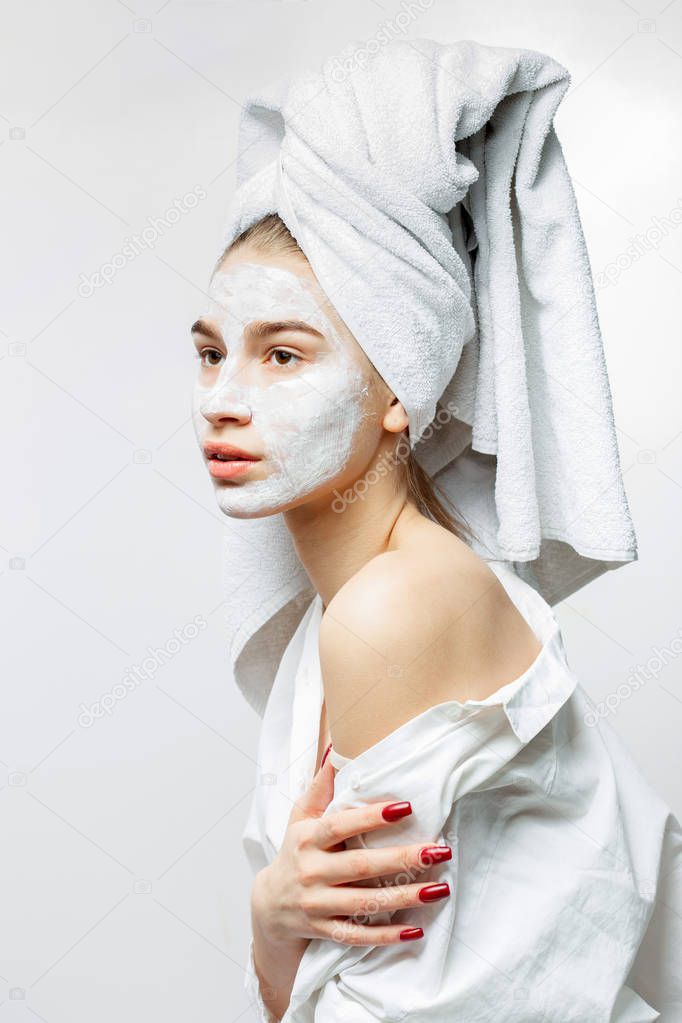 Beautiful young woman dressed in white clothes with a white towel on her hair and cosmetic mask on her face holds her hand on her naked shoulder