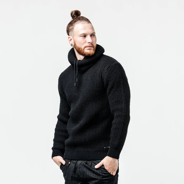 Elegante hombre con barba y peinado de moño vestido con suéter de punto negro y pantalones posa en el estudio sobre el fondo blanco — Foto de Stock