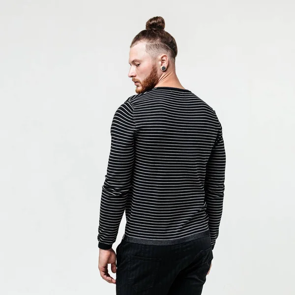 Stylish young man with beard and bun hairstyle wearing a dark striped jumper and trousers poses poses in the studio on the white background — Stock Photo, Image