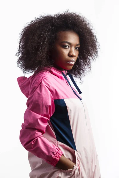 Saucy young curly brown-haired girl dressed in the pink sport jacket stands at the white background in the studio — Stock Photo, Image