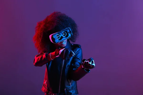 Curly dark haired girl dressed in black leather jacket and gloves uses the virtual reality glasses on her head in the dark studio with neon light