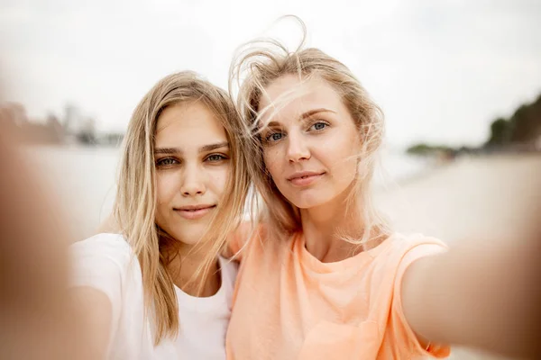 Duas jovens lindas garotas loiras tiram uma selfie na praia em um dia quente de vento — Fotografia de Stock