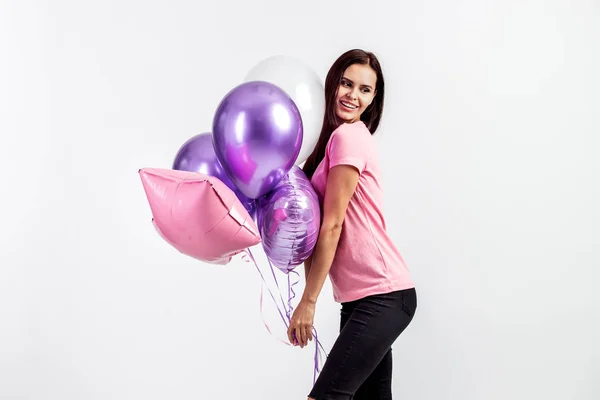 Linda chica sonriente vestida con camiseta rosa y jeans sostiene globos en sus manos sobre el fondo blanco en el estudio — Foto de Stock