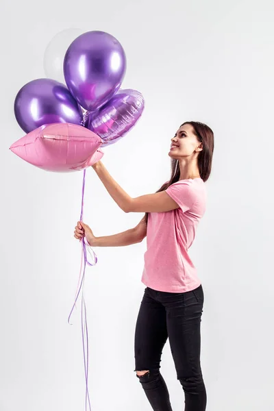 Linda chica sonriente vestida con camiseta rosa y jeans sostiene globos en sus manos sobre el fondo blanco en el estudio — Foto de Stock