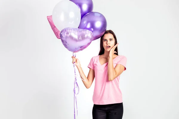 Menina surpresa vestida com camiseta rosa e jeans segura balões em suas mãos no fundo branco no estúdio — Fotografia de Stock