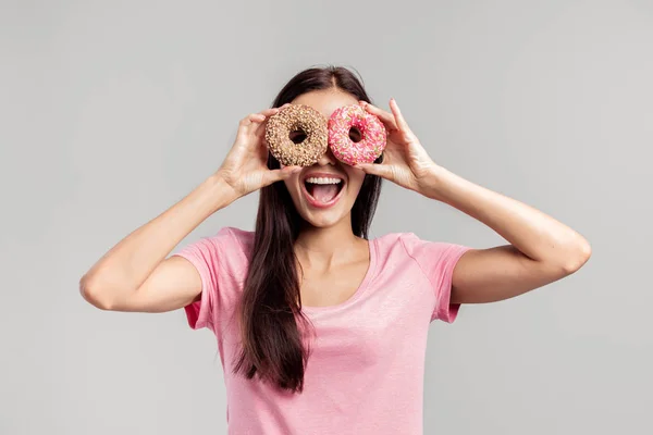 Забавная девушка в розовой футболке держит два ярких пончика возле глаз, как очки на белом фоне. — стоковое фото