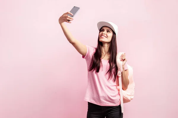 Hermosa chica en gorra blanca y con una mochila en los hombros vestida con camiseta rosa y vaqueros negros está haciendo selfie en el fondo rosa en el estudio — Foto de Stock