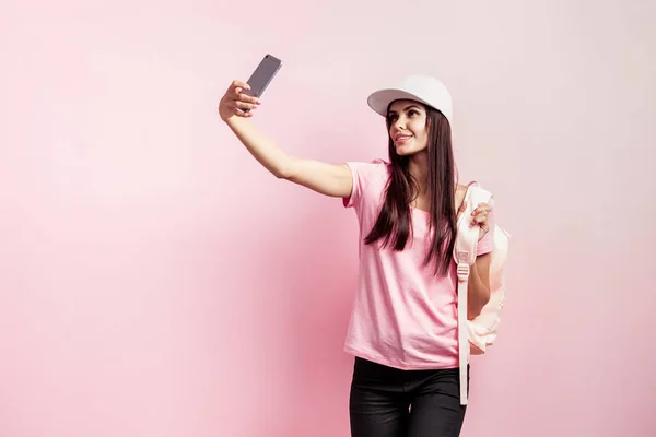 Hermosa chica en gorra blanca y con una mochila en los hombros vestida con camiseta rosa y vaqueros negros está haciendo selfie en el fondo rosa en el estudio — Foto de Stock