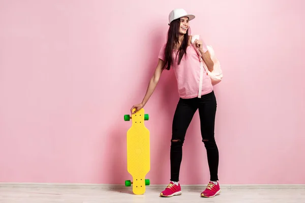 Hermosa chica en gorra blanca y con una mochila en el hombro vestida con camiseta rosa y pantalones vaqueros negros con monopatín amarillo en el fondo rosa en el estudio — Foto de Stock