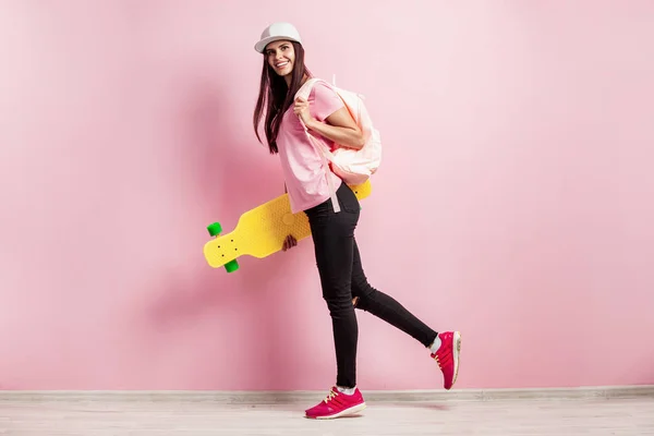 Elegante ragazza in berretto bianco e con uno zaino sulla spalla vestito di t-shirt rosa e jeans neri stand con skateboard giallo sullo sfondo rosa in studio — Foto Stock