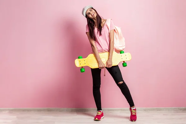 Elegante chica en gorra blanca y con una mochila en su hombro vestida con camiseta rosa y jeans negros se levanta con monopatín amarillo sobre el fondo rosa en el estudio — Foto de Stock