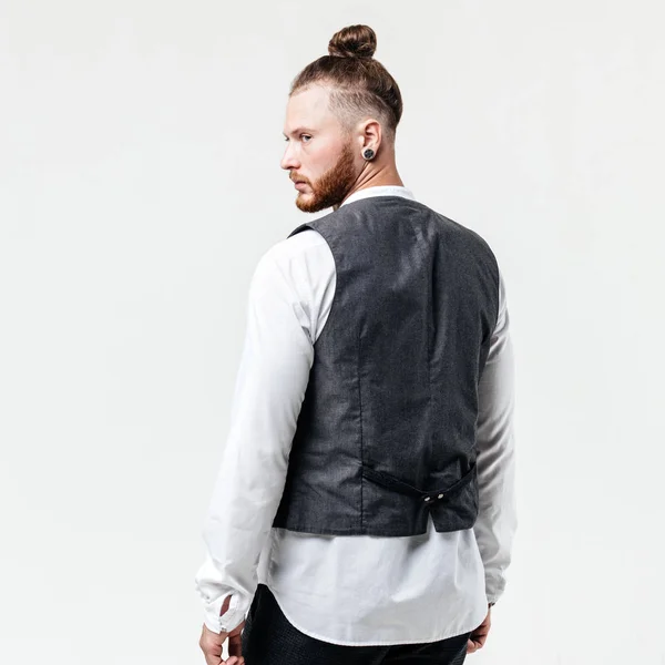 Bel homme élégant avec barbe et un chignon coiffure vêtue d'un gilet gris sur une chemise blanche et pantalons gris pose dans le studio sur le fond blanc — Photo