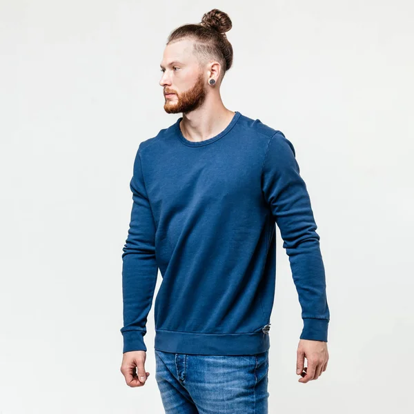 Homem elegante com barba e penteado de pão vestido de camisola de manga comprida azul e jeans posa no estúdio no fundo branco — Fotografia de Stock