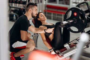 Siyah spor kıyafetlerini giymiş ince esmer kız bacaklarını bir antrenör gözetiminde spor salonunda ile ağırlık kaldırma