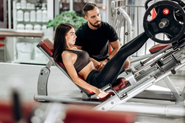İnce kız siyah spor kıyafetlerini giymiş ile bacaklarını bir antrenör gözetiminde spor salonunda ağırlık kaldırma