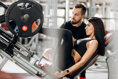 İnce kız siyah spor kıyafetlerini giymiş ile bacaklarını bir antrenör gözetiminde spor salonunda ağırlık kaldırma