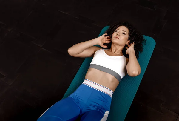 Slim kudrnaté tmavé vlasy dívka oblečená v sportovní oblečení dělat cvičení pro tisk na podložce pro fitness v tělocvičně — Stock fotografie
