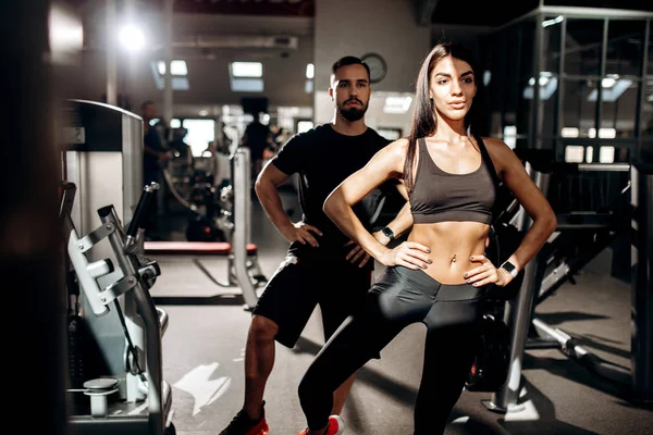 Menina de cabelos escuros magro vestido com roupas esportivas pretas e treinador de fitness estão fazendo aquecimento no ginásio — Fotografia de Stock