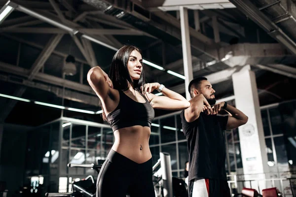 Menina de cabelos escuros magro vestido com roupas esportivas pretas e treinador de fitness estão fazendo aquecimento no ginásio — Fotografia de Stock