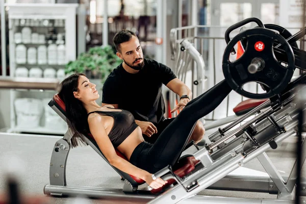 İnce kız siyah spor kıyafetlerini giymiş ile bacaklarını bir antrenör gözetiminde spor salonunda ağırlık kaldırma