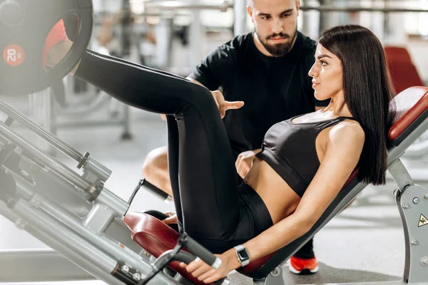 Atletisk flicka klädd i svart sportkläder lyfta vikter med benen i gymmet under överinseende av en coach — Stockfoto