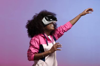 Modern kıvırcık kahverengi saçlı kız pembe spor ceket giymiş Studio neon arka plan üzerinde sanal gerçeklik gözlük kullanır