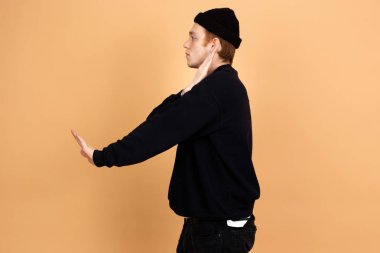 Siyah kot pantolon, kazak ve şapka giymiş adam dansçı stüdyoda bej renkli arka plan üzerinde elleriyle hareketleri yapıyor