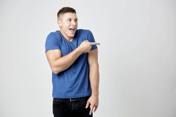 Cheerful guy dressed in blue t-shirt points his finger to the side — Stock Photo, Image