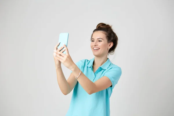 Chica alegre vestida con una camiseta azul claro sonríe y mantiene el teléfono móvil en su mano — Foto de Stock