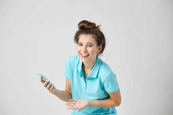 Chica alegre vestida con una camiseta azul claro sonríe y mantiene el teléfono móvil en su mano —  Fotos de Stock