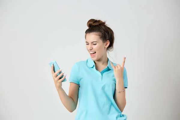 Glad flicka klädd i ljus blå t-shirt leenden håller mobiltelefon i handen och visar rock tecken — Stockfoto