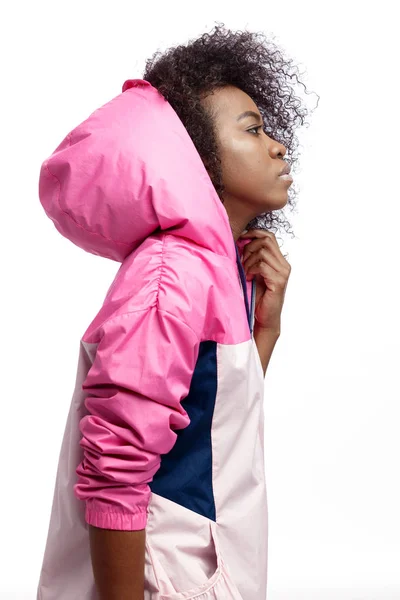 Mod young curly brown-haired girl dressed in the pink hooded sports jacket poses at the white background in the studio — Stock Photo, Image