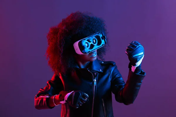 Lockiges dunkelhaariges Mädchen in schwarzer Lederjacke und Handschuhen nutzt die Virtual-Reality-Brille auf dem Kopf im dunklen Studio mit Neonlicht — Stockfoto