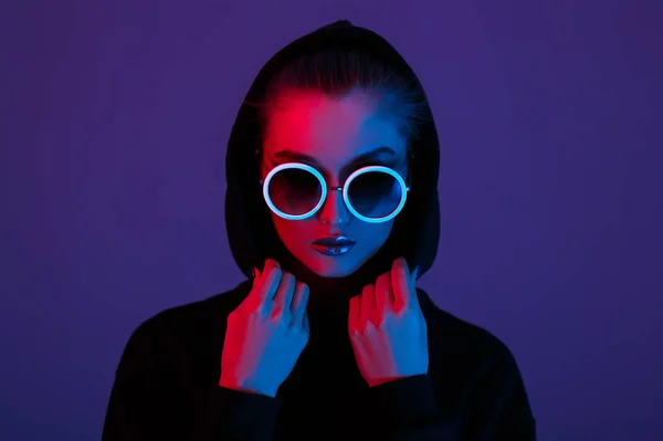 Modemädchen im schwarzen Pullover mit Kapuze und runder Sonnenbrille in rotem und blauem Neonlicht im Atelier — Stockfoto