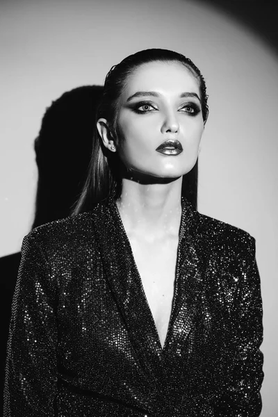 Retrato preto e branco da menina da moda com cabelo puxado para trás e maquiagem elegante em um vestido brilhante standin g ao lado da parede em holofotes brilhantes — Fotografia de Stock