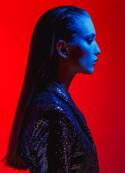 Móda dívka s dlouhými vlasy a stylový make-up v černé lesklé šaty pózy na červeném pozadí v neonové světlo v ateliéru — Stock fotografie