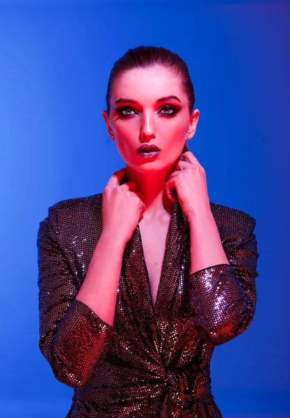 Módní dívka s dlouhými vlasy a stylový make-up v černé lesklé šaty pózy na modrém pozadí v neonové světlo v ateliéru — Stock fotografie