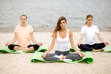 Üç ince genç kız oturup bir yoga sıcak bir günde paspaslar Nehri yanındaki kum plajındaki gözünü kapatma ile pozlar