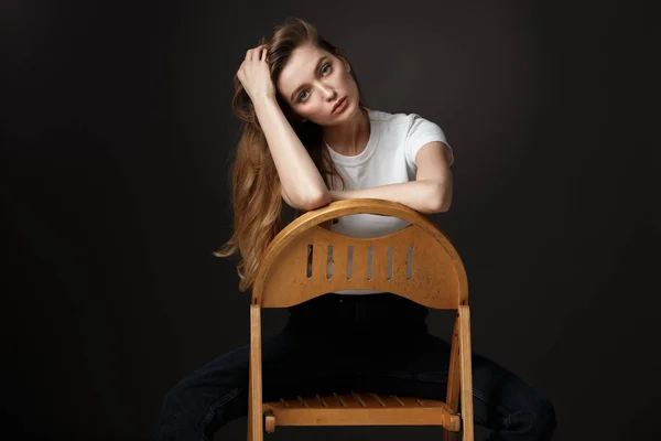 Beautiful brunette girl with long flowing hair dressed in white t-shirt and jeans poses sitting on the wooden chair on the black background in the studio — Stock Photo, Image