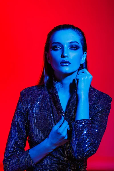 Móda dívka s dlouhými vlasy a stylový make-up v černé lesklé šaty pózy na červeném pozadí v neonové světlo v ateliéru — Stock fotografie