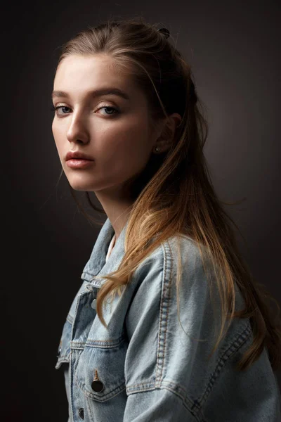 Portrait of beautiful brunette girl with hair tied back dressed in jeans jacket on the dark background in the studio — Stock Photo, Image