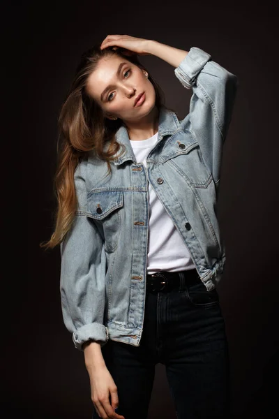 Gorgeous brunette girl with long flowing hair dressed in jeans jacket and jeans poses standing on the dark background in the studio — Stock Photo, Image