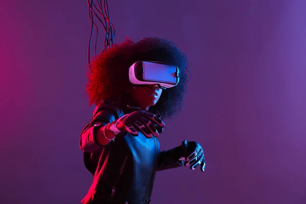 Mod chica de pelo oscuro rizado vestido con chaqueta de cuero negro y guantes utiliza las gafas de realidad virtual en su cabeza en el estudio oscuro con luz de neón — Foto de Stock