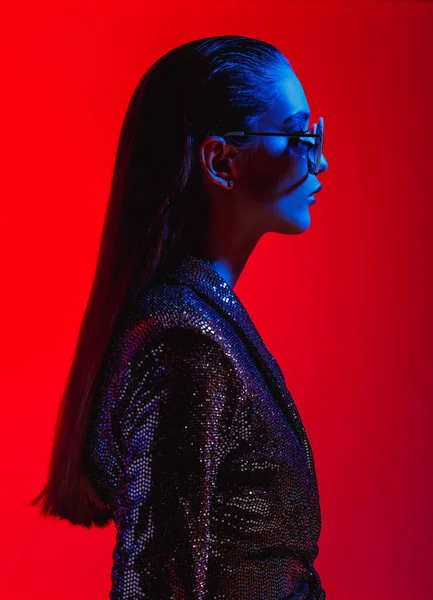 Módní dívka s dlouhými vlasy a kulatými slunečními brýlemi v černých lesklých šatech pózuje v neonovém světle ve studiu — Stock fotografie
