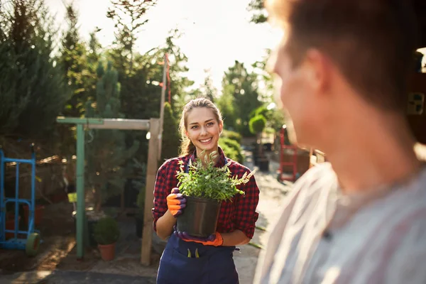 Mooie lachende meisje tuinman houdt pot met planten en kijken naar de man in de prachtige tuin op een zonnige dag — Stockfoto