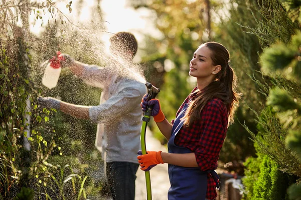 Trabaja en el jardín. Chica jardinero rocía agua y un chico rocía fertilizante en las plantas en el hermoso vivero-jardín en un día soleado . — Foto de Stock